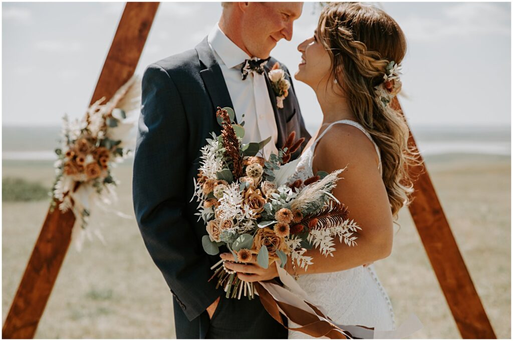 wedding couple portrait holding wedding florals in front of a ceremony arch with flowers.